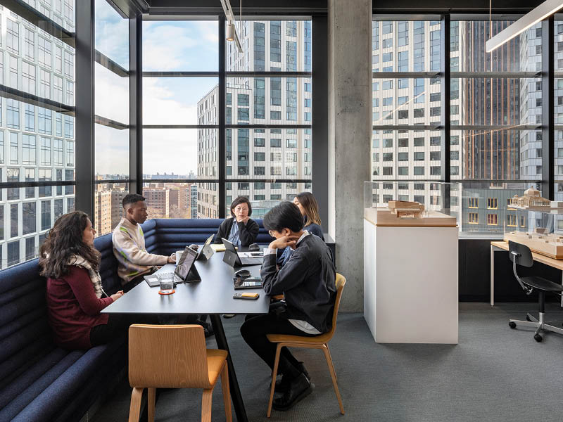 (c) Magda Biernat. Photograph of four people sitting at a table in front of large windows with city views.