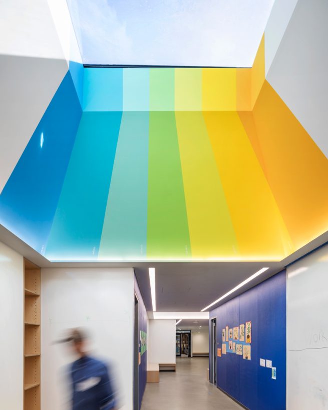 teacher walks down sawtooth hallway below a colorful striped skylight and streak of light