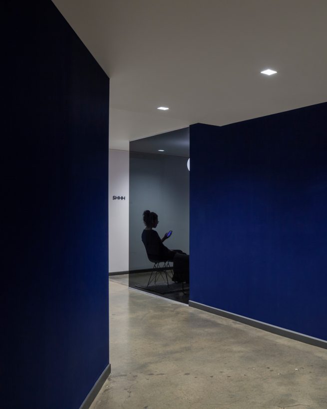 woman sits behind glass wall at the end of a blue walled shortcut and a graphic that reads SHHH