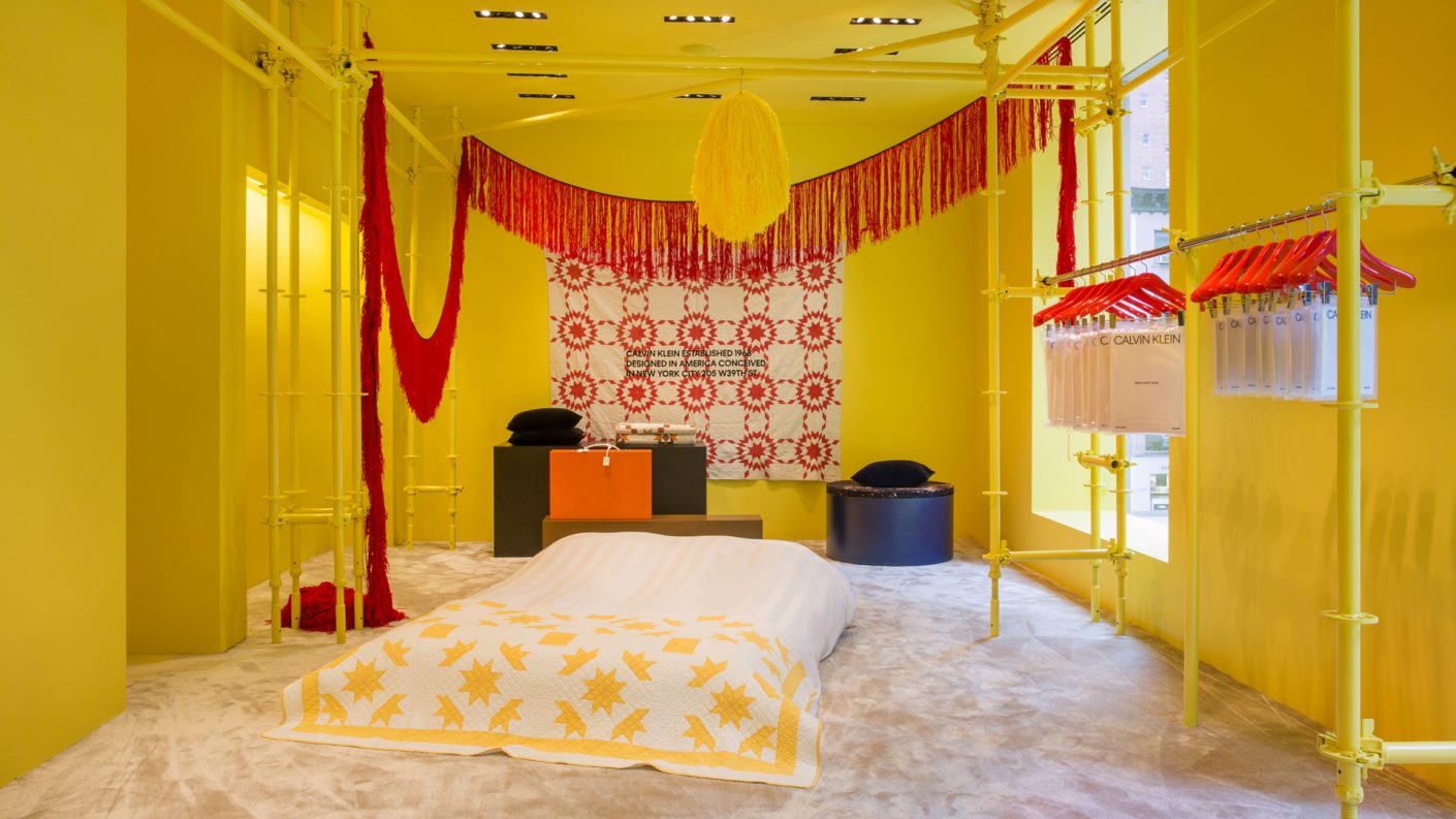 bed in center of a yellow room with red hanging art displays and red and yellow quilts