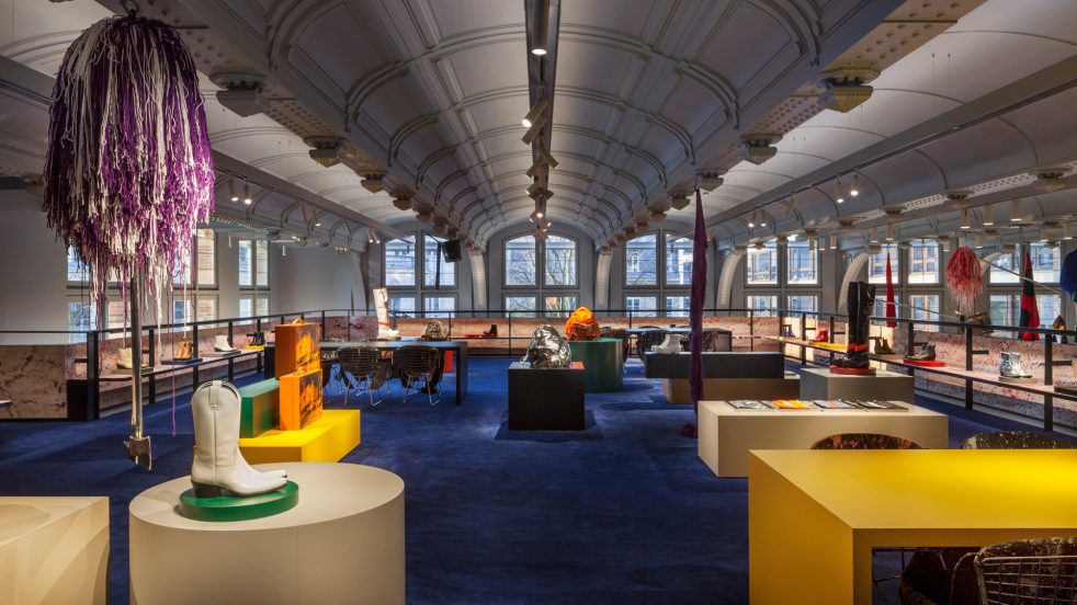 shoe displayed on custom formica pedestals with Sterling Ruby mobiles between vaulted ceilings and a deep blue carpet in mezzanine level