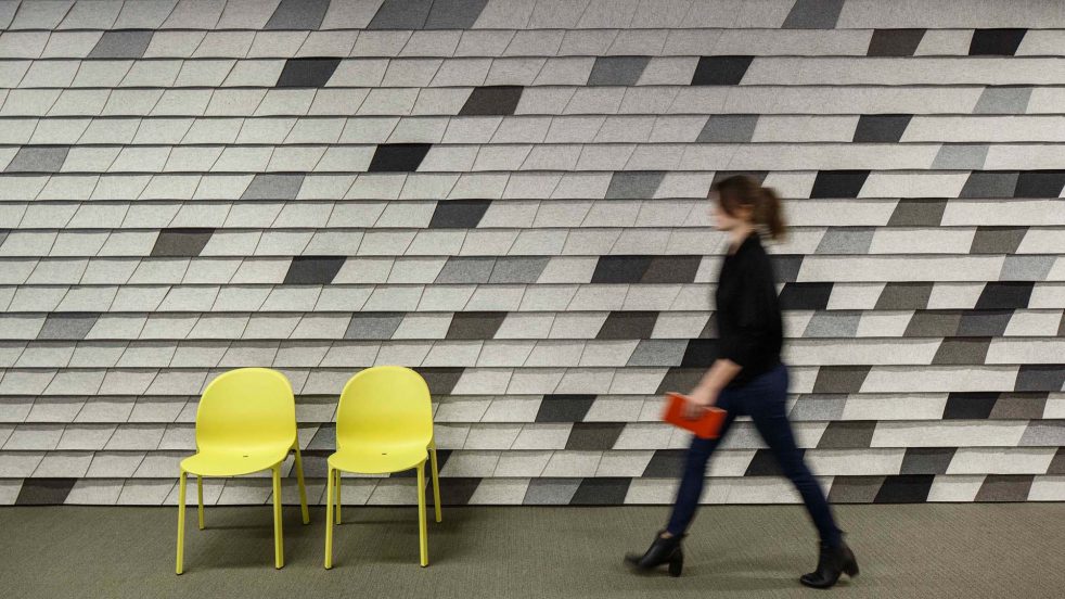 woman passes in front of ARO Shingle mounted in shades of grey on wall, and two yellow chairs