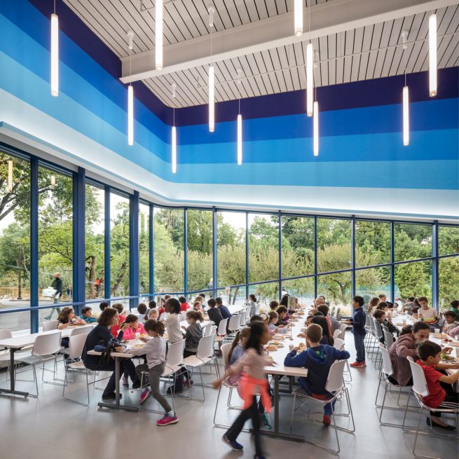 primary school students eating lunch in the cafeteria / student center with blue striped wall graphic and large windows overlooking the campus