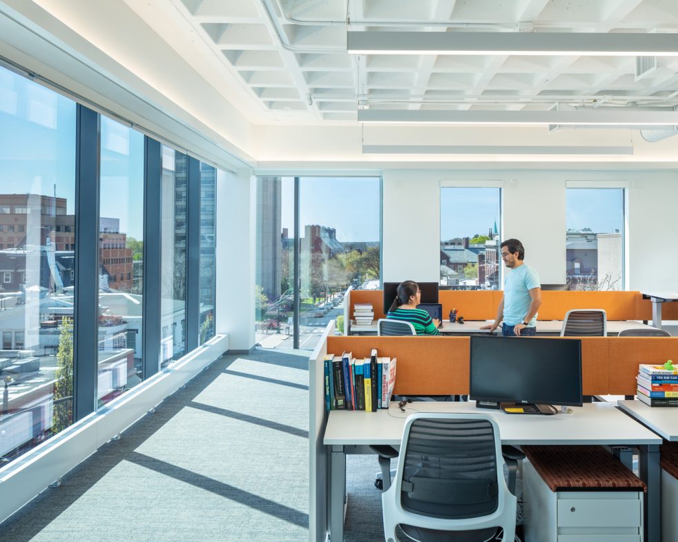 Two people talking in a corner workspace with wall of windows