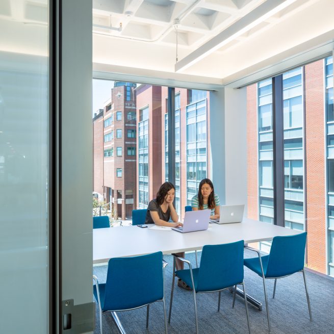 Two students working on laptops in corner glass meeting room