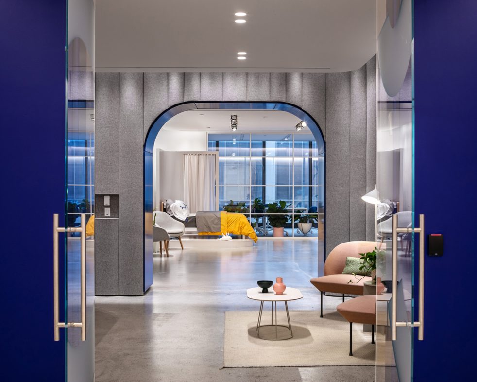 Two open glass doors look towards reception seating area, a display bed, and glass conference room beyond