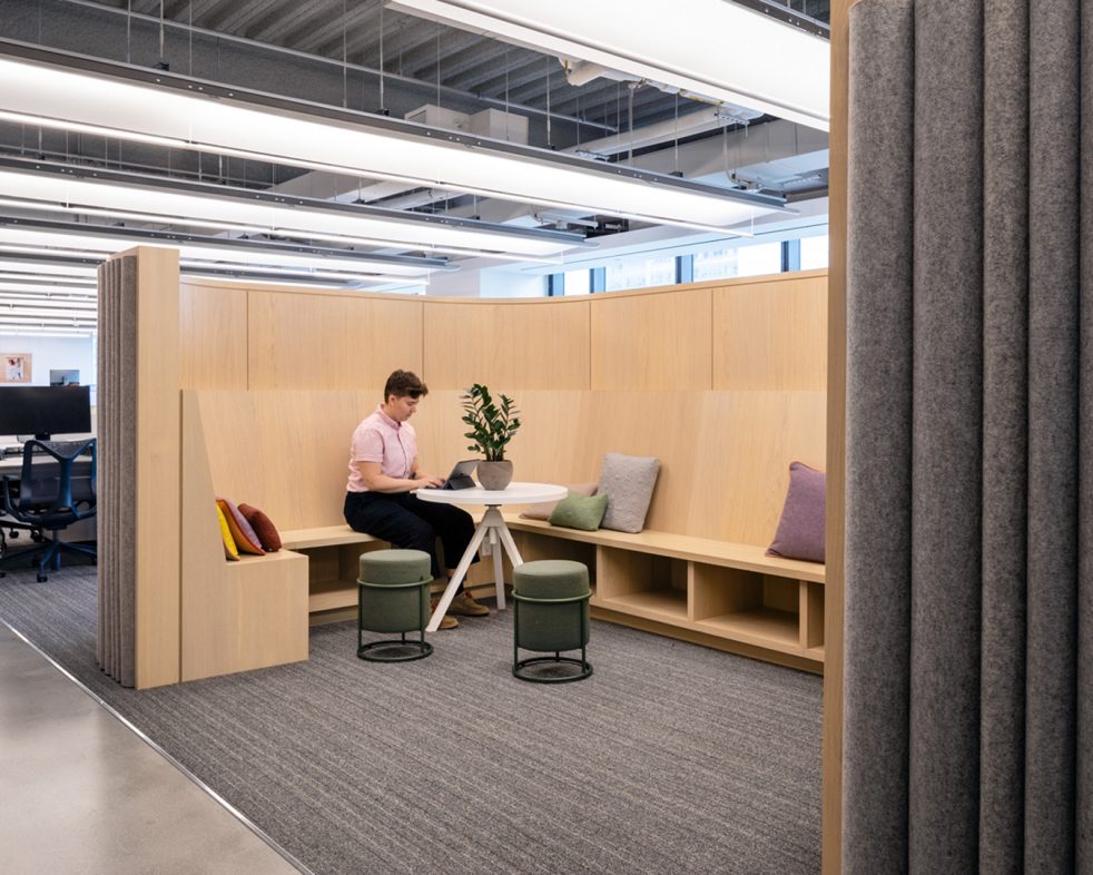 Person working on a laptop in a work pod with a wooden bench and surrounded by grey felt