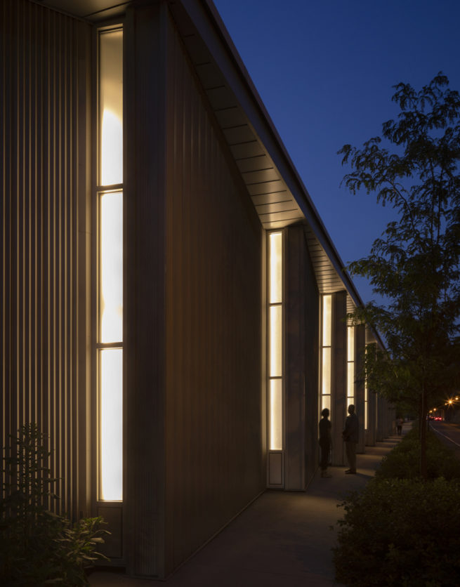 Two people stand by angled light panels along the building façade at night