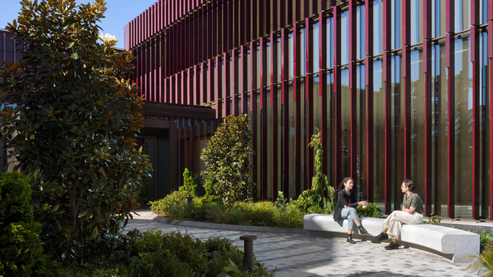 Two people sit on a stone bench in front a burgundy, terra cotta-clad building. Image Credit: Rafael Gamo