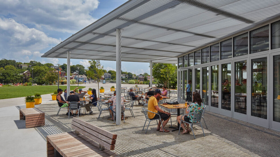 People sit under a pavilion's shaded canopy on a sunny day.