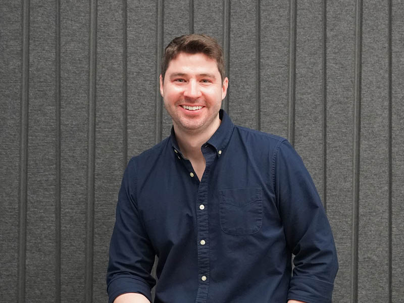 Man in a blue shirt sits against a gray backdrop.