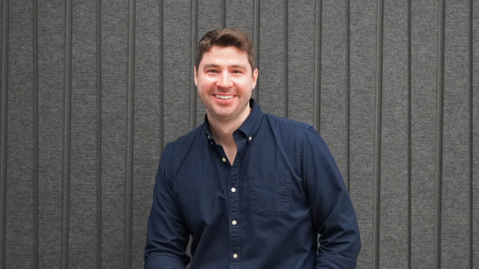 Man in a blue shirt sits against a gray backdrop.