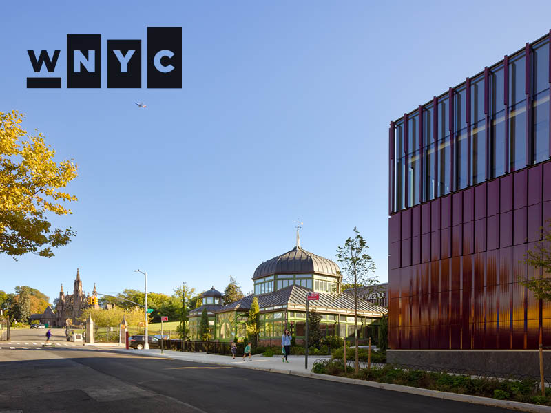 Parent and child walk in front of a historic greenhouse, with historic cemetery gates in the background and new burgundy, terra cotta-clad visitor's center in the foreground